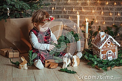 Cute little girl is sitting by the Christmas tree on the floor in the room. Near the baby is a gingerbread house and a basket with Stock Photo