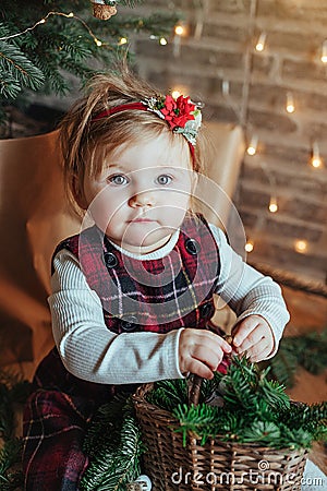 Cute little girl is sitting by the Christmas tree on the floor in the room. Near the baby is a gingerbread house and a basket with Stock Photo