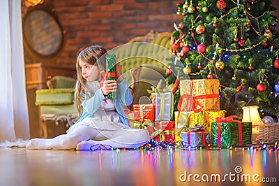 Cute little girl sitting on the floor in the room near the festive Christmas tree, holding a gift box Stock Photo