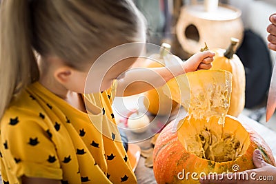 Cute little girl sitting on kitchen table, helping her father to carve large pumpkin, smiling. Halloween family lifestyle. Stock Photo