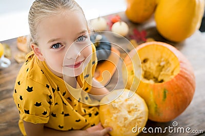 Cute little girl sitting on kitchen table, helping to carve large pumpkin, looking at camera and smiling. Halloween. Stock Photo