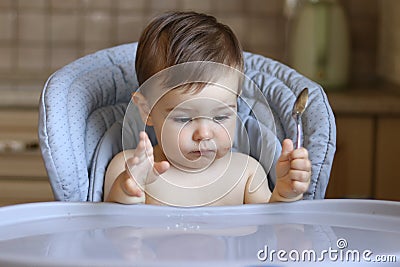 Cute little hungry baby boy holds spoon in his hand and looks at empty table waiting for food Stock Photo