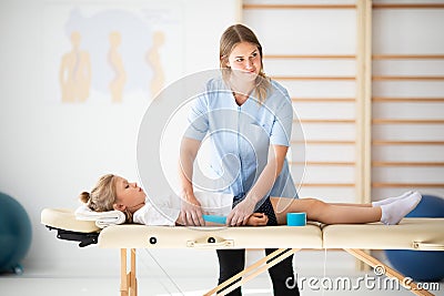 Little injure girl lying on massage table and exercising with young female doctor Stock Photo