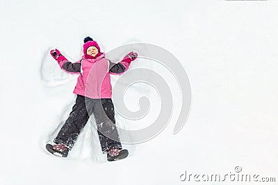 Cute little kid girl in warm ski sport suit making snow angel outdoors. Kid having fun lying on snowdrift after snow storm in Stock Photo