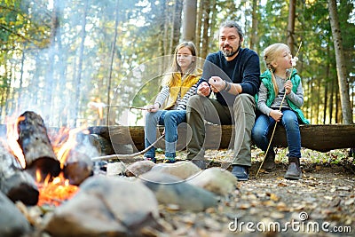 Cute little sisters and their father roasting marshmallows on sticks at bonfire. Children having fun at camp fire. Camping with ki Stock Photo