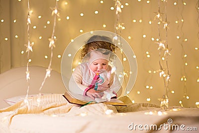 Cute little toddler girl reading book in dark room with Christmas lights Stock Photo