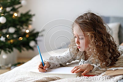Cute lovely curly little female child lies on comfortable bed in her bedroom with decorated fir tree in background Stock Photo