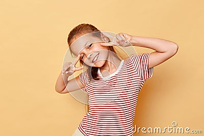 Cute lovely sweet charming dark haired little girl wearing striped t-shirt showing v-sign near eyes smiling and winking to camera Stock Photo