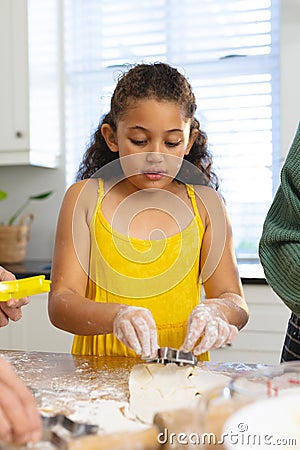 Cute multiracial girl with messy hands cutting dough with pastry cutter on table in kitchen Stock Photo