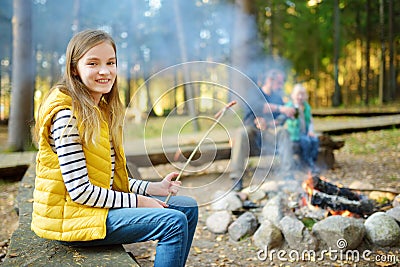 Cute preteen girl roasting hotdog on a stick at bonfire. Child having fun at camp fire. Camping with kids in fall forest. Stock Photo