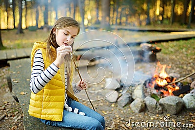 Cute preteen girl roasting marshmallows on stick at bonfire. Child having fun at camp fire. Camping with children in fall forest. Stock Photo