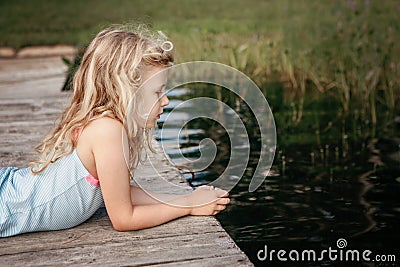 Cute pretty Caucasian blonde girl lying on wooden dock pier by lake river and looking in water. Pensive sad thinking child kid Stock Photo