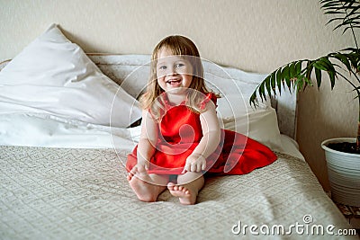 Cute red-haired blue-eyed emotional baby girl laughs. Lies on the bed in the bedroom, the bed is a white bed, a red dress for a Stock Photo