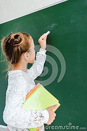cute schoolkid holding chalk and books near green chalkboard . Stock Photo