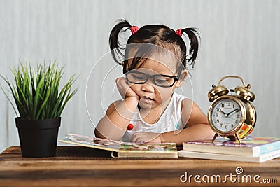 Cute serious looking little asian toddler wearing spectacle reading a book on a table Stock Photo