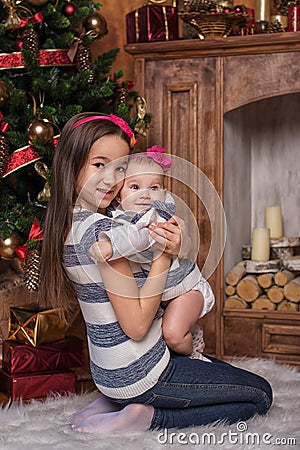 Cute sisters sitting on white carpet near christmas tree and fireplace, wearing striped sweaters and red headbands. Smiling toddle Stock Photo