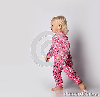 Laughing barefooted blonde baby kid girl in pink warm comfortable jumpsuit is running aside over grey wall background Stock Photo