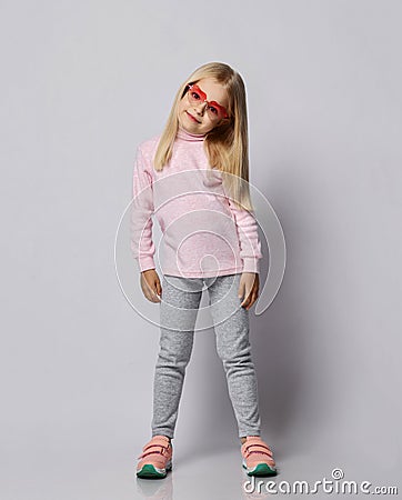 Cute blonde kid girl in stylish smoked heart-shaped sunglasses, turtleneck shirt sweater and pants is posing over gray Stock Photo