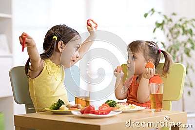 Cute smiling child and toddler girls playing and eating spaghetti with vegetables for healthy lunch sitting in a white sunny kitch Stock Photo