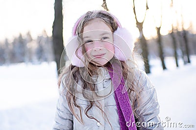 Cute smiling kid girl 5-6 year old wear scarf and fluffy headphones over sunny light in snowy cold park close up. Winter season. Stock Photo