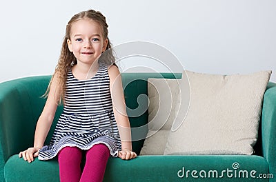 Cute smiling preschooler girl wearing stripy navy blue dress sitting on a couch. Stock Photo