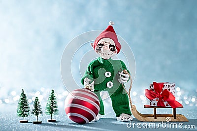 Cute Smiling Santas Helper Elf Holding Christmas Bauble and Pulling Christmas Gift on a Sledge. North Pole Christmas Scene. Stock Photo