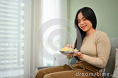 Cute smiling young Asian woman sitting on couch while eating a piece of cake. People, food and domestic life Stock Photo