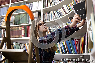 Pretty student stands on a stepladder in the library to find the right book. Woman in public library Stock Photo