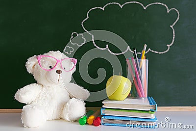 Cute teddy bear wearing glasses next to a pile of books and an apple in a classroom. Drawn thought bubble on a blackboard Stock Photo