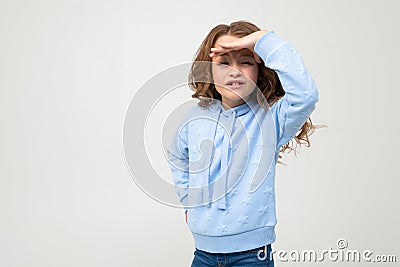 Cute teenage girl in a blue hoodie seeks out while holding a hand near her forehead on a light gray background Stock Photo