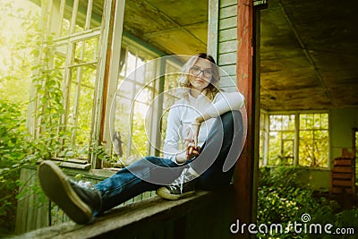 A cute teenager with blonde hair and jeans is sitting on the windowsill of a wooden abandoned house on a sunny summer day Stock Photo