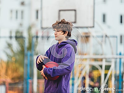 Cute teenager in violet hoodie playing basketball. Young boy with ball learning dribble and shooting on the city court. Hobby Stock Photo