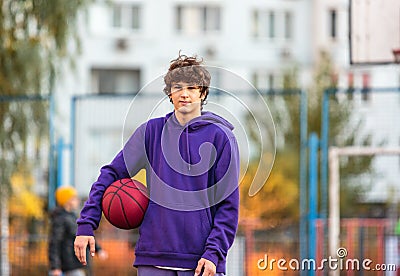 Cute teenager in violet hoodie playing basketball. Young boy with ball learning dribble and shooting on the city court. Hobby Stock Photo