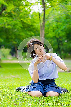 Cute Thai schoolgirl is sitting on the grass and doing heart symbol. Stock Photo