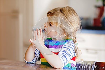 Cute toddler girl praying to God at home. Child using hands for pray and thank for food. Christian tradition. Stock Photo