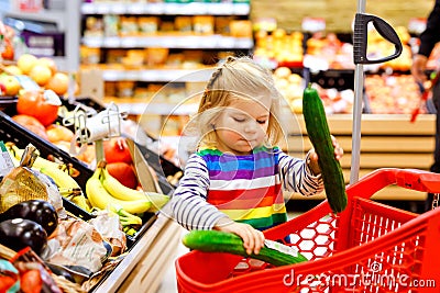 Cute todler girl pushing shopping cart in supermarket. Little child buying fruits. Kid grocery shopping. Adorable baby Stock Photo