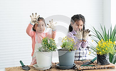 Cute two Asian little girls smiling looking at dirty hands in gloves and apron while having fun planting tree and change flowers Stock Photo