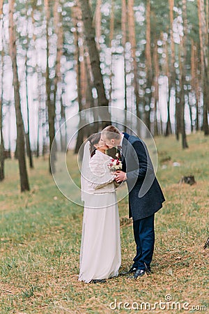 Cute white dressed bride with her handsome groom kissing in green pine forest Stock Photo