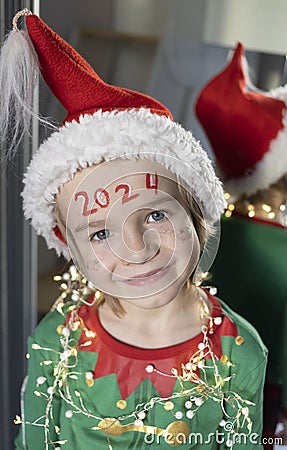 cute 6 year old boy in elf costume, santa hat, garland beads, New Year 2024 numbers written on forehead Stock Photo