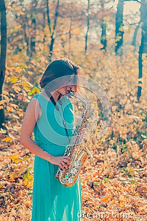 Cute young brunette girl in a long blue dress plays the yellow saxophone in the autumn forest Stock Photo