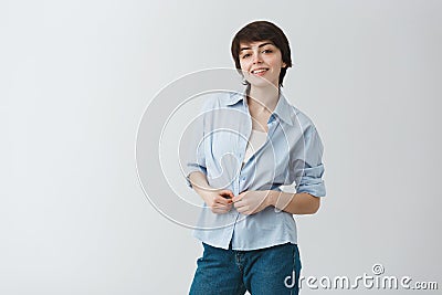 Cute young female student with short dark hair brightfully smiling, buttoning up shirt and looking in camera with happy Stock Photo