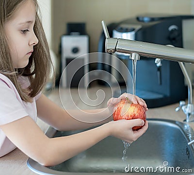 Cute young girl washing and sanitizing fresh fruit. Wash food. Child hands holding tasty apple under running water in kitchen sink Stock Photo