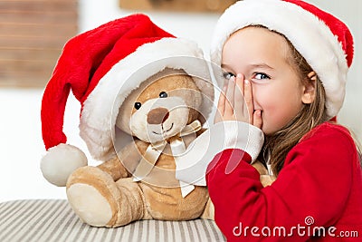 Cute young girl wearing santa hat whispering a secret to her teddy bear christmas present toy. Cheeky kid with teddy bear. Stock Photo