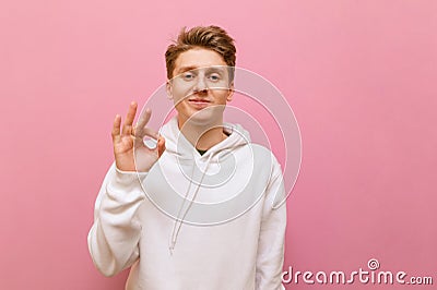 Cute young man in white casual clothes looks into the camera with a smile on his face and shows a hand gesture OK. Handsome blond Stock Photo