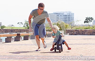 Dad has his back while he rides his bike. A happy toddler boy riding is toy motorbike outside while his father runs Stock Photo