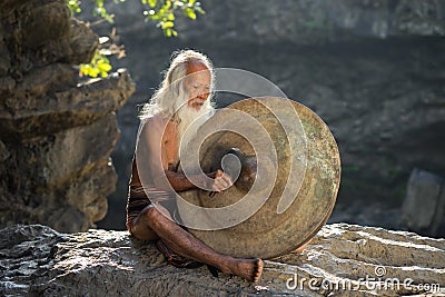 Daklak, Vietnam - Mar 9, 2017: Portrait of minority man in traditional dress knocking a copper gong in forest in Buon Me Thuot Editorial Stock Photo