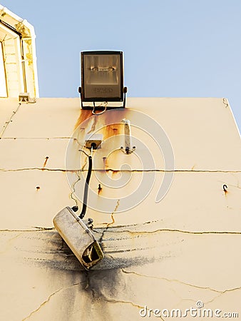 Damaged CCTV camera hanging on a cable on yellow wall, old flood light above the camera. Front element is broken and there is sign Stock Photo