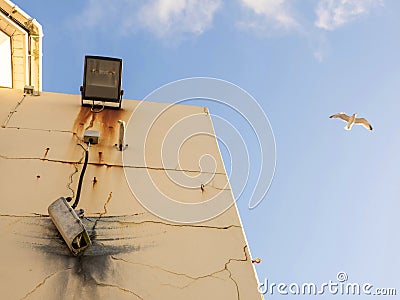 Damaged CCTV camera hanging on a cable on yellow wall, old flood light above the camera. Front element is broken and there is sign Stock Photo