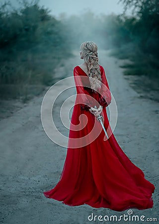 Dangerous blonde queen in red fashion lush dress Hides a dagger behind. Backdrop dark fantasy forest in fog. Concept Stock Photo