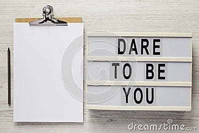 `Dare to be you` on a lightbox, cliboard with blank sheet of paper on a white wooden background, top view. Flat lay, overhead, Stock Photo